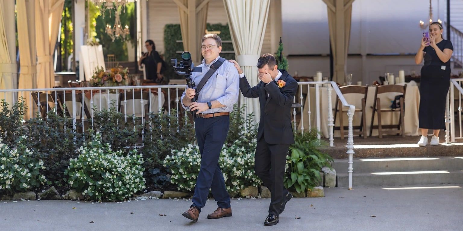 Brian, camera rig in hand, walks a groom to his first look while the groom covers his eyes and keeps a hand on Brian's shoulder