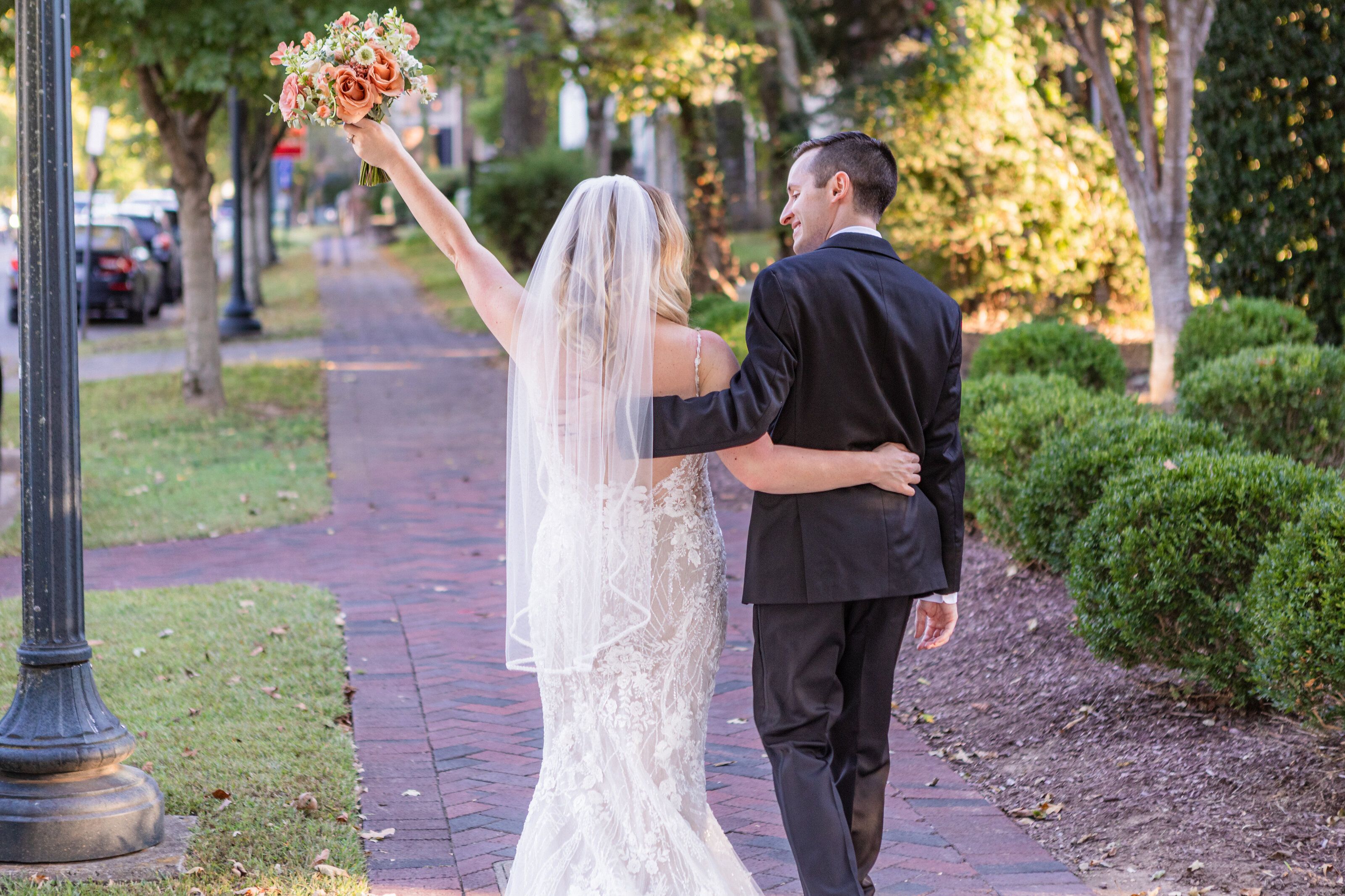 A couple walks arm in arm down a brick sidewalk, one raising a bouquet in the air