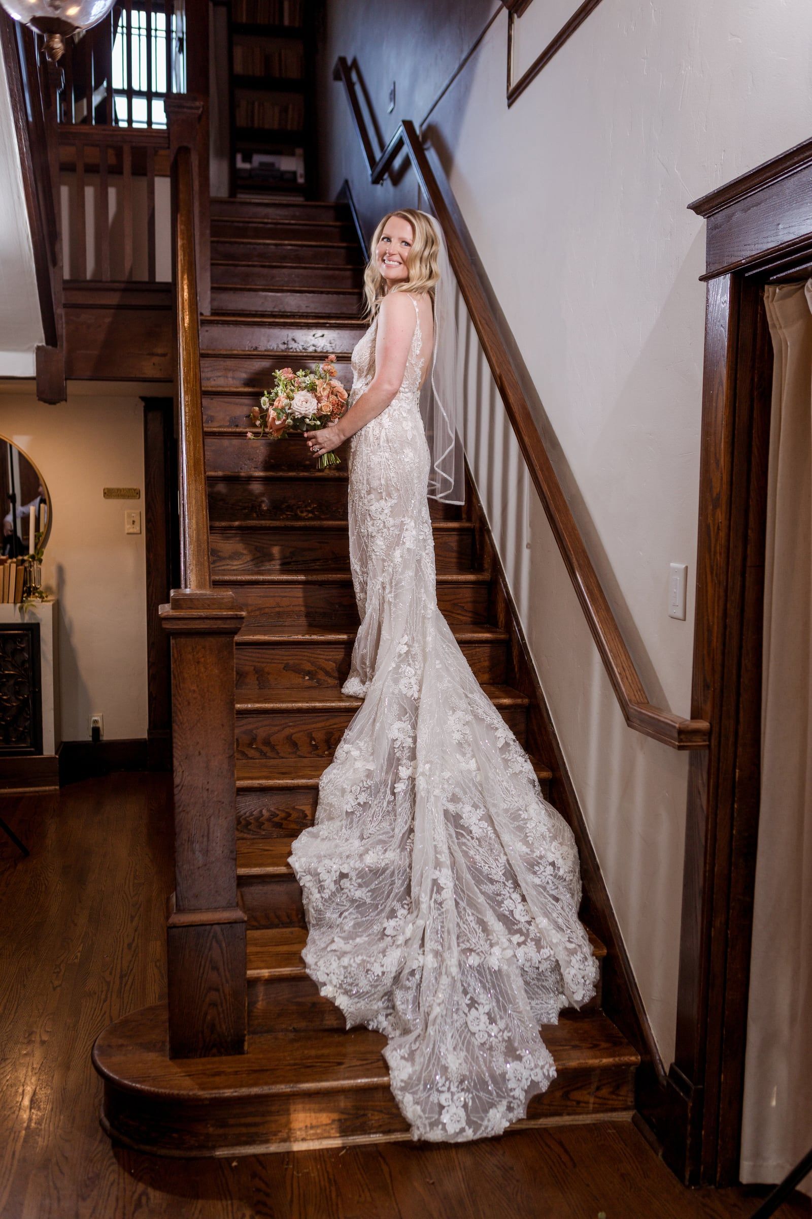 Caitlin pauses halfway down the staircase, her lace train spilling over the steps