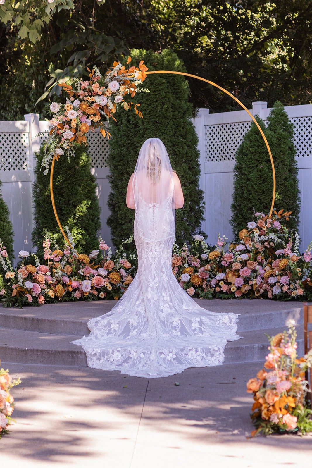 Caitlin from behind at the gold ceremony arch, her cathedral train fanned across the stone