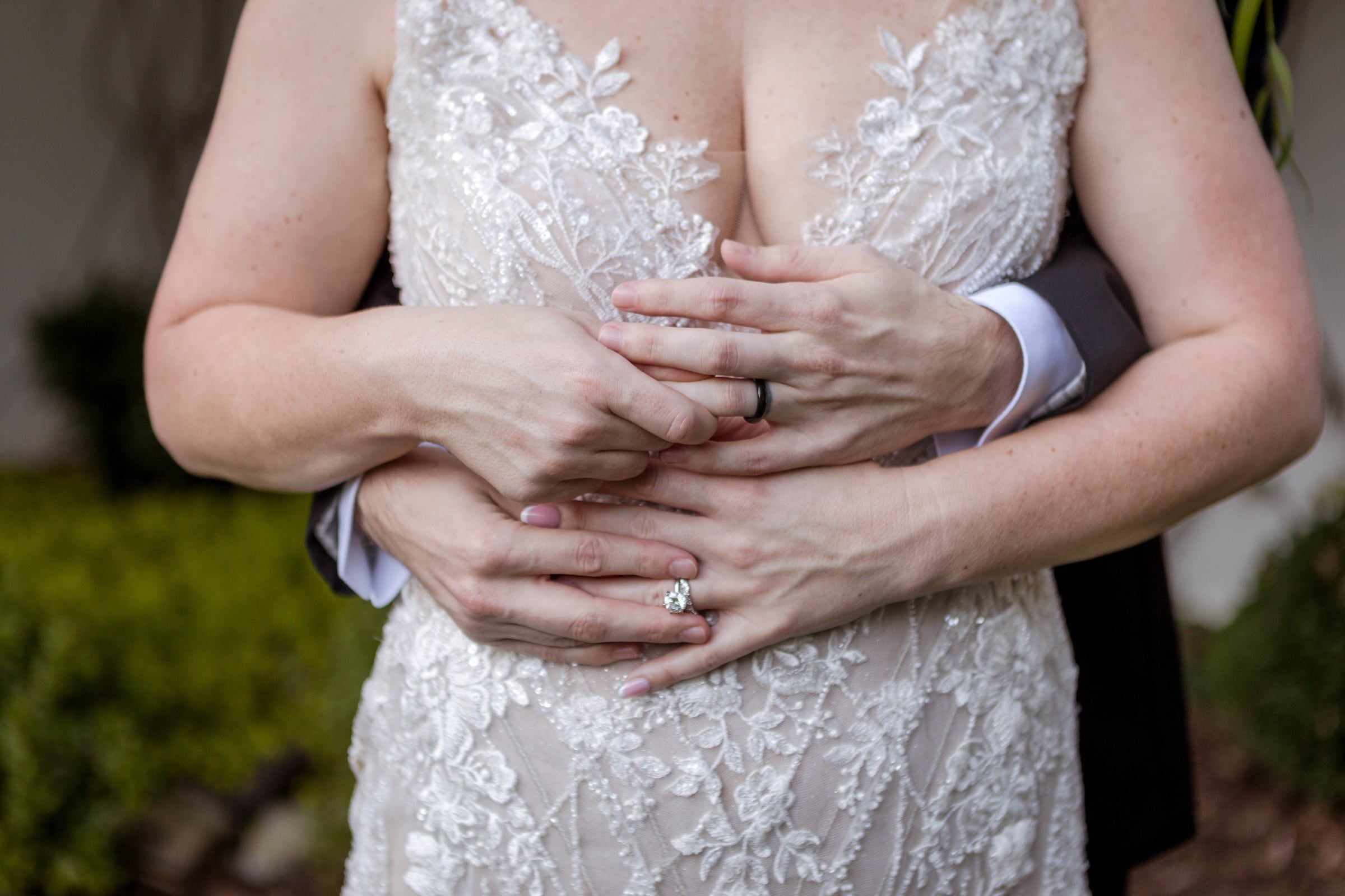 Hands wrapped around Caitlin's waist, her engagement ring catching the light against her lace gown