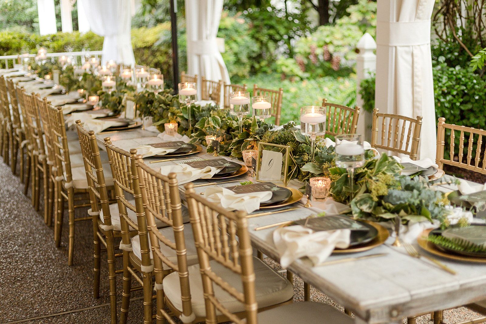 A long reception table set with candles, greenery and gold chairs under a white tent