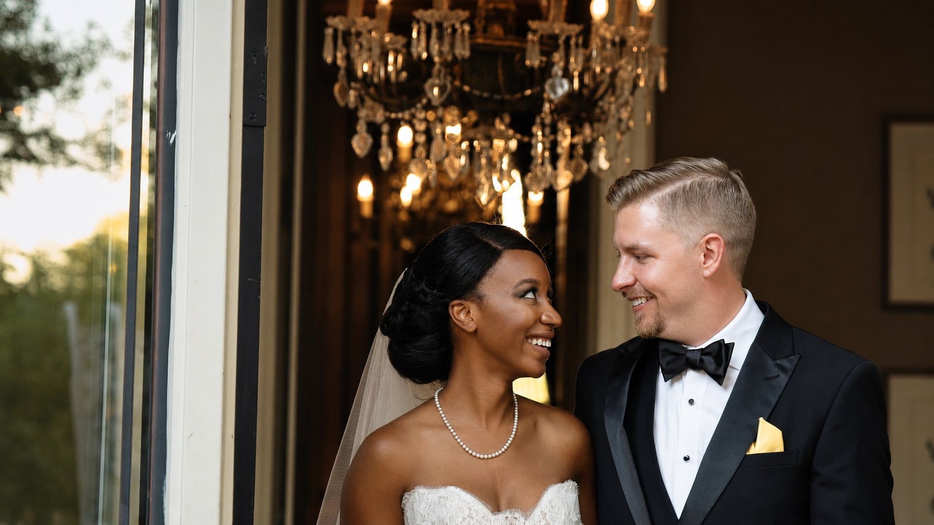 Jocasta and Nick smile at each other in a doorway at East Ivy Mansion, a crystal chandelier glowing behind them