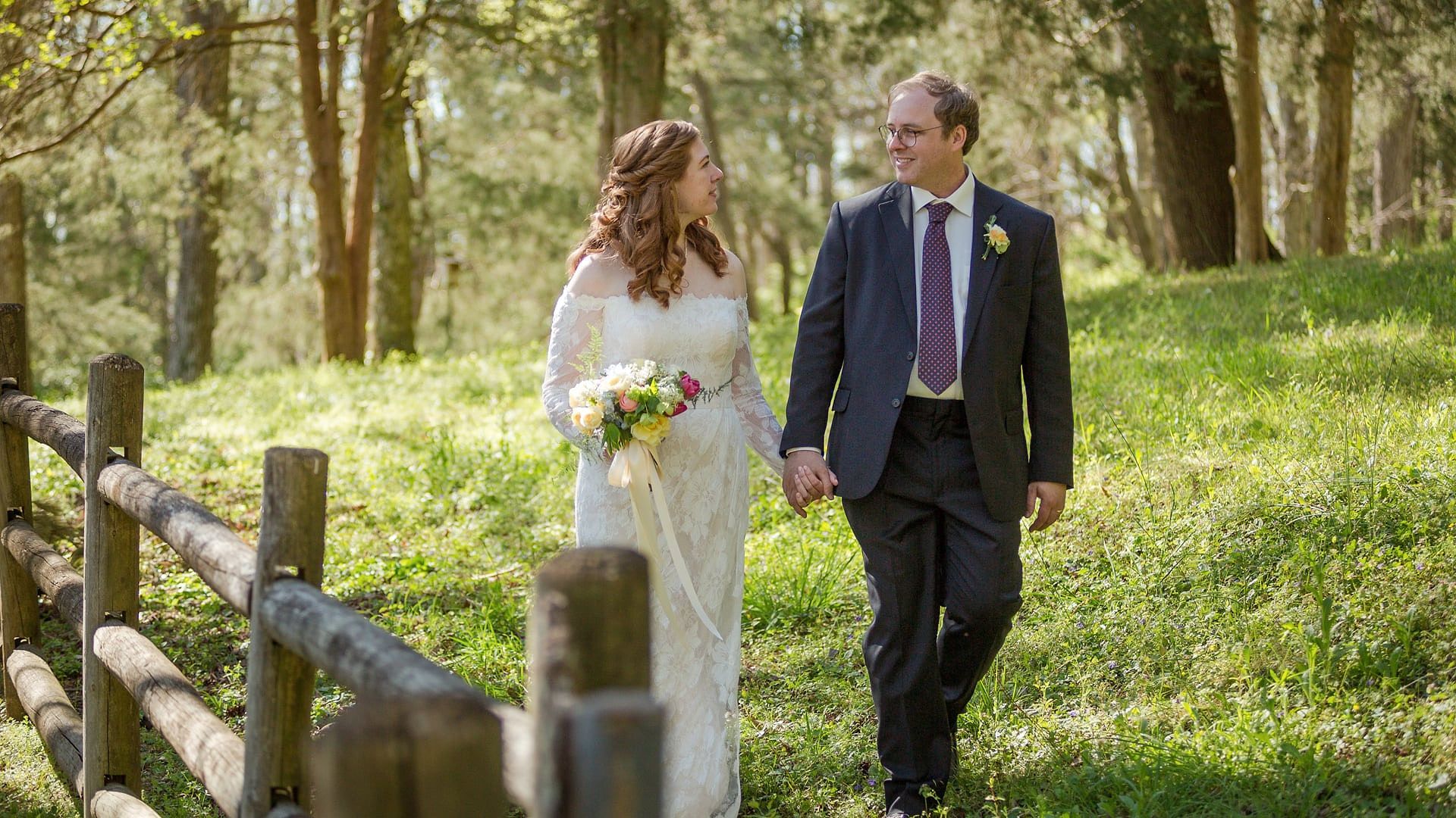 Mamie and Will walk hand in hand along a split-rail fence on a green hillside, smiling at each other