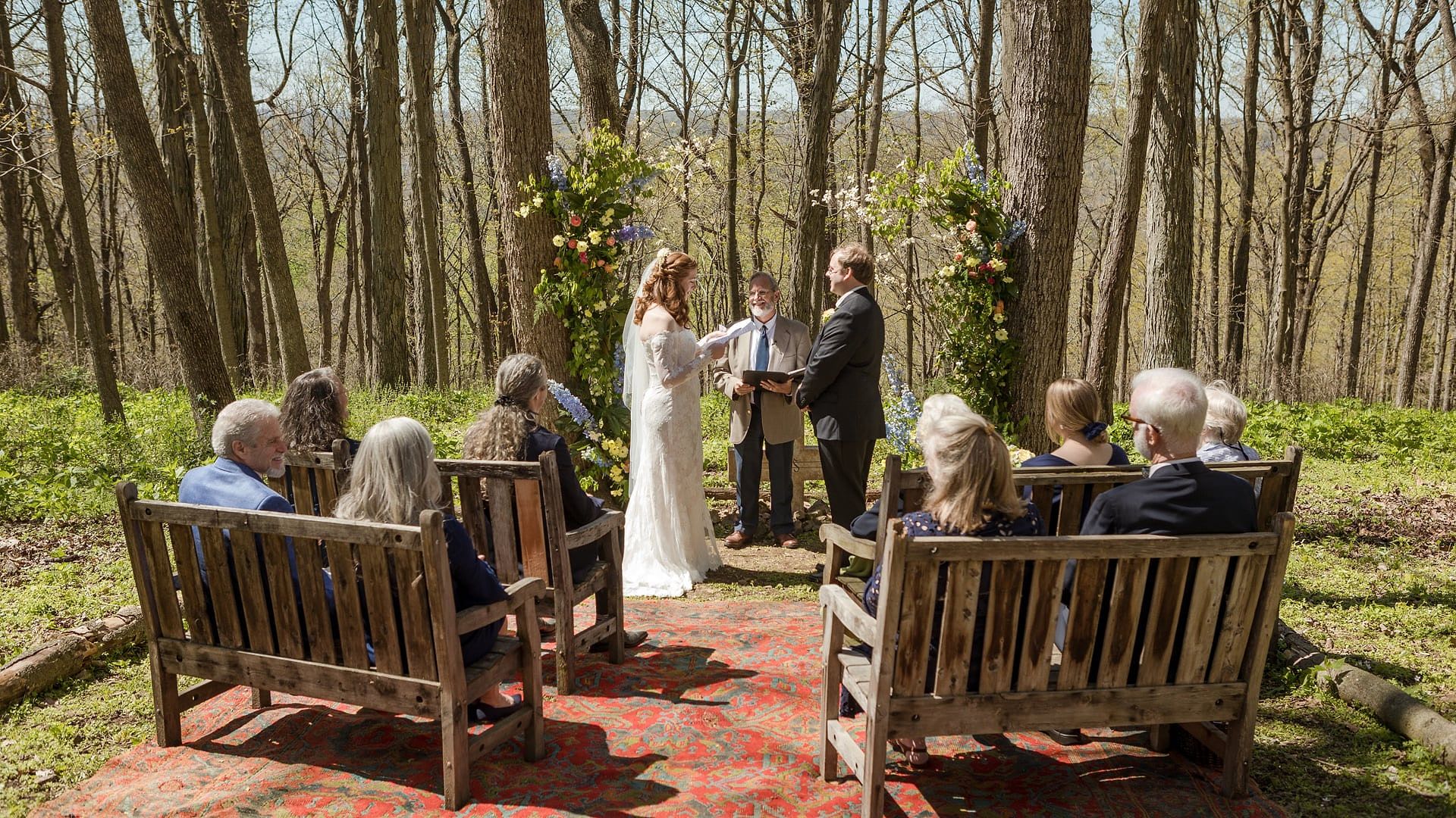 Mamie and Will stand before the officiant under a flowered arch in the woods as family watches from wooden benches
