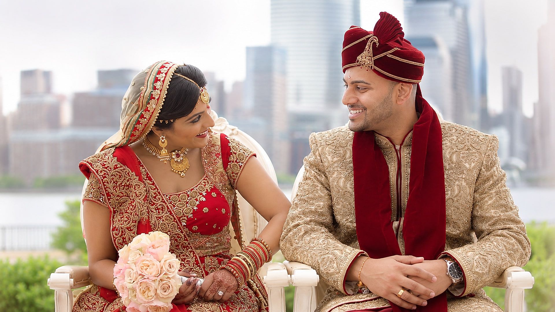 Preksha and Ankit, in red and gold wedding attire, smile at each other with the Manhattan skyline behind them