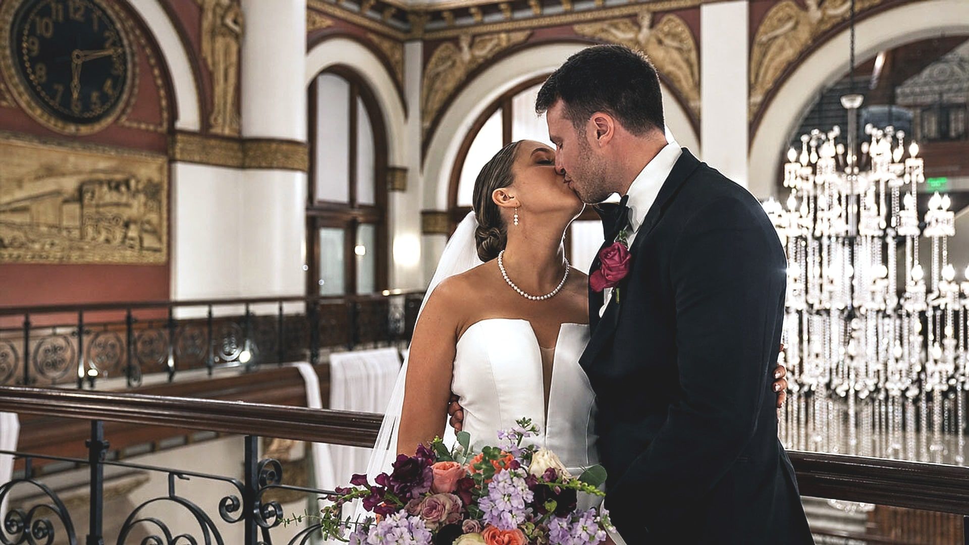 Stephanie and Jacob kiss on the balcony at Union Station Hotel, with the station's clock and chandeliers behind them