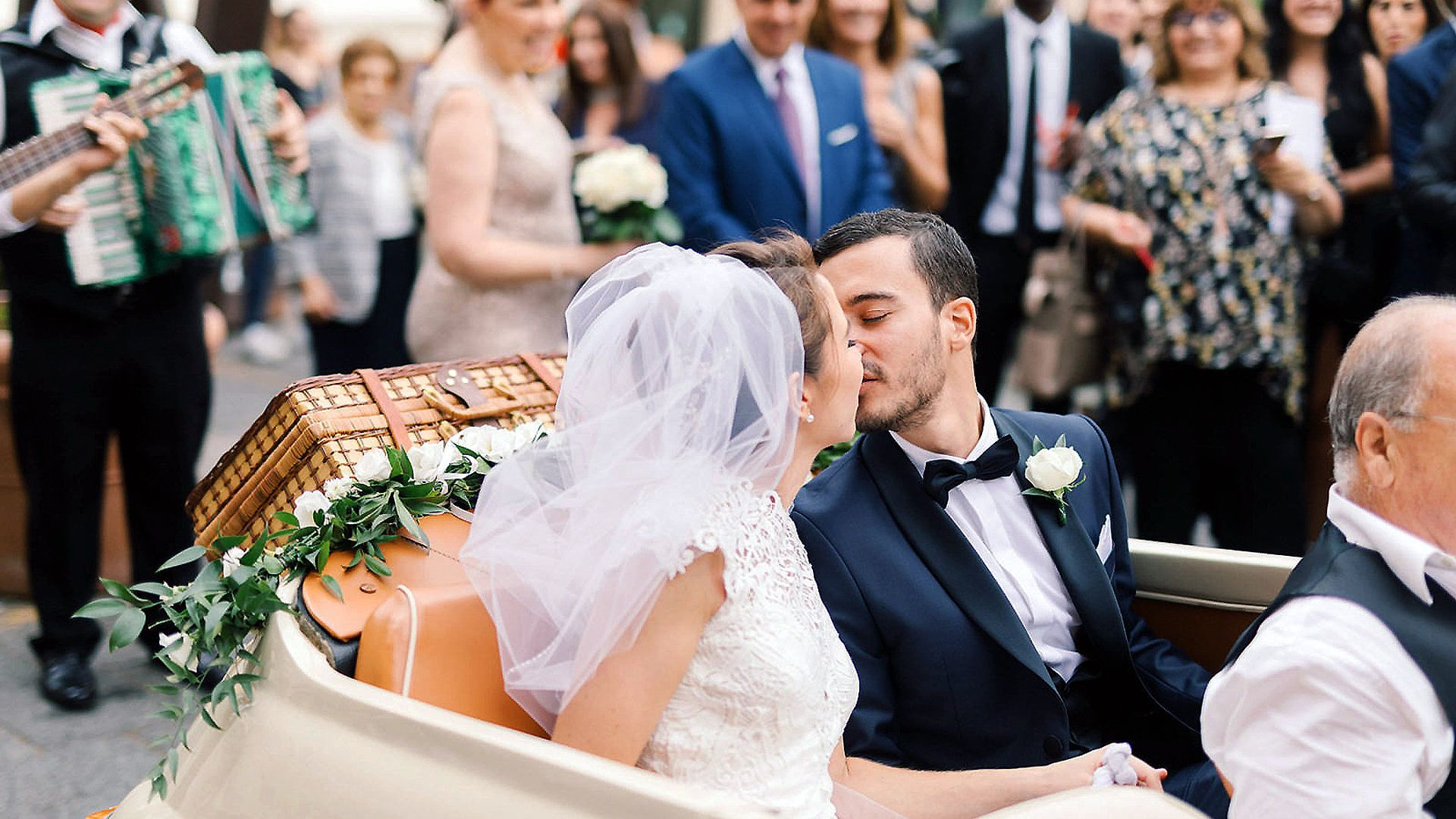 Veronica and Massimo kiss in the back of an open vintage car as guests and an accordion player celebrate around them