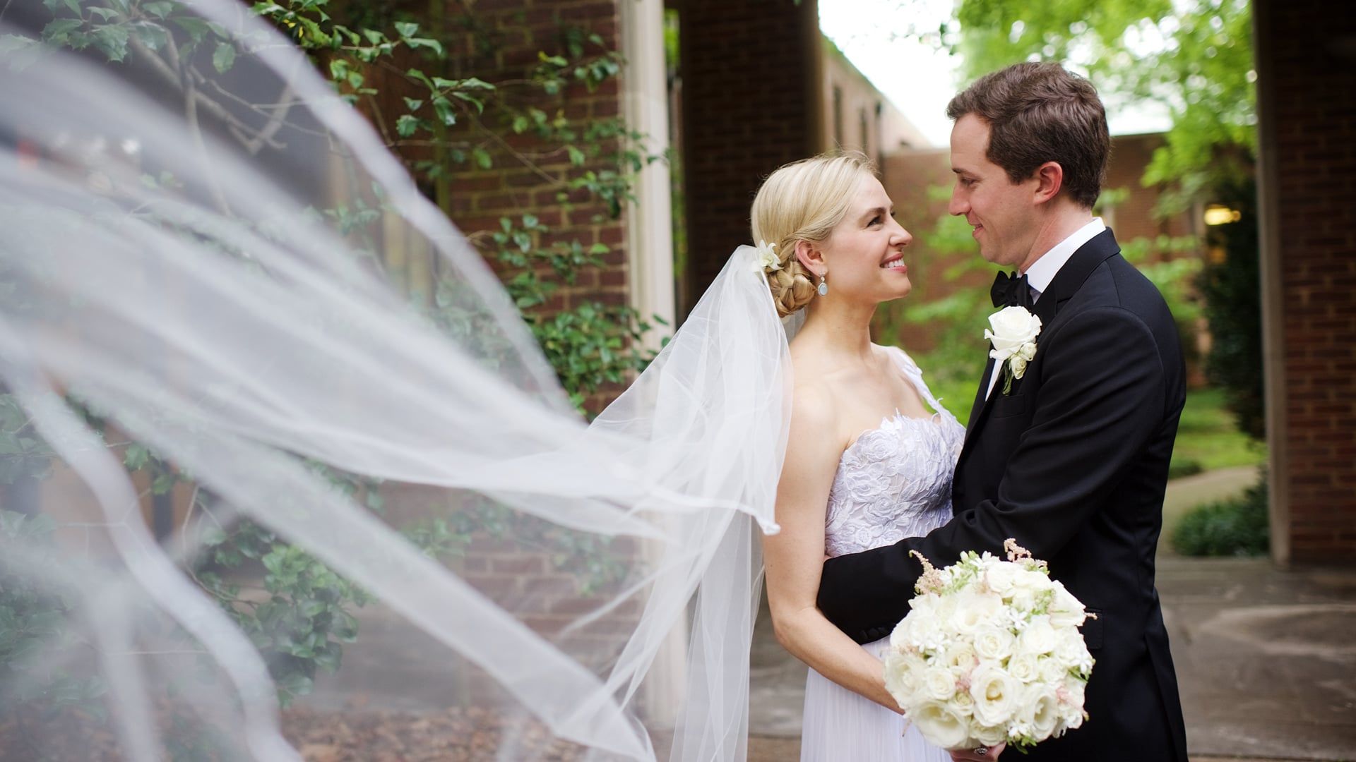 Frances and Jim smile at each other as her veil blows across the frame outside a brick building