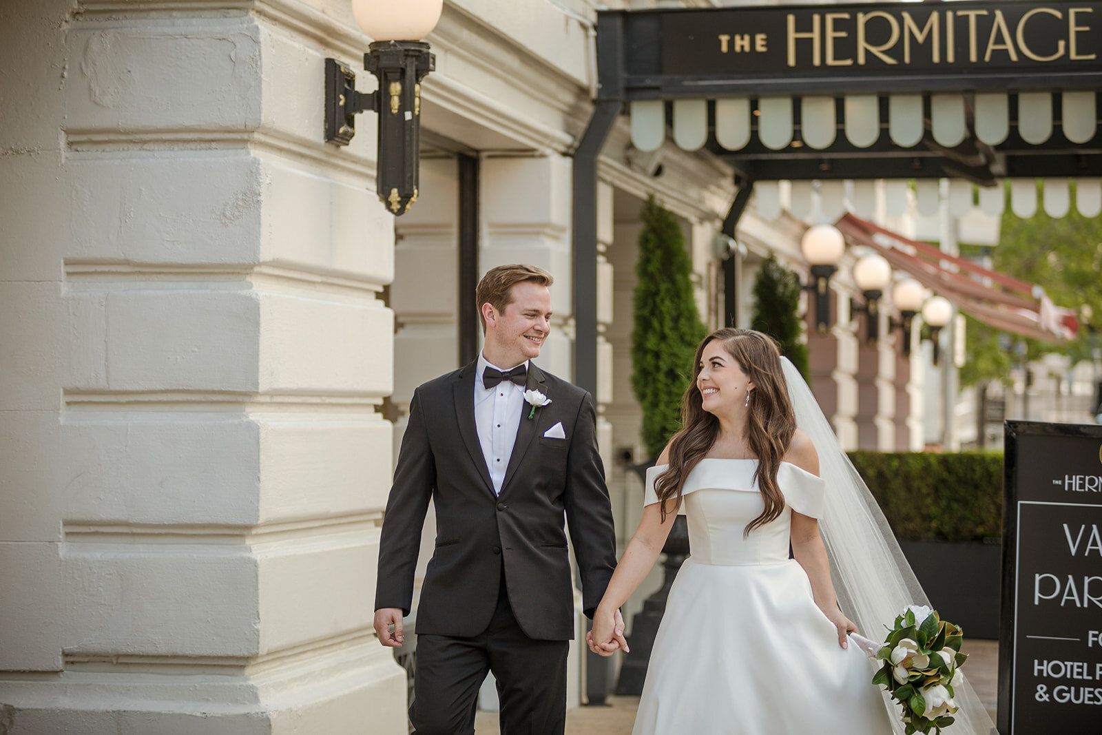 Shelby and Reid walk hand in hand past the entrance of The Hermitage Hotel, smiling at each other