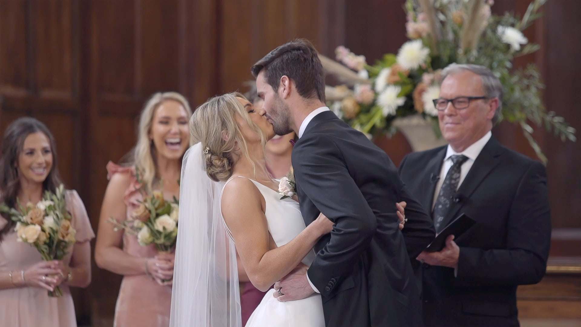 A couple shares their first kiss at the altar as their wedding party laughs behind them