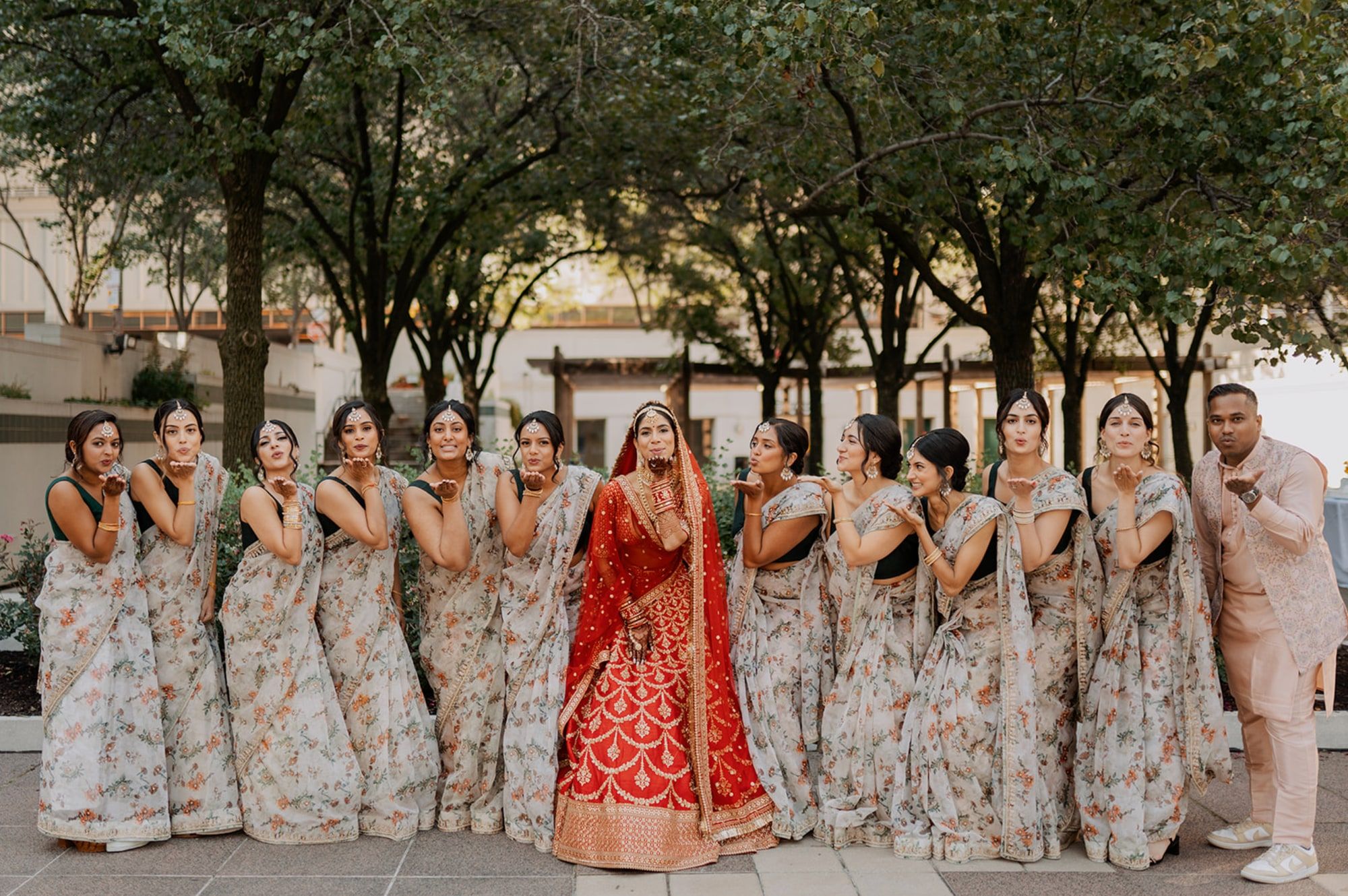 Bride in a red lehenga blowing kisses to the camera with her bridesmaids in matching floral saris