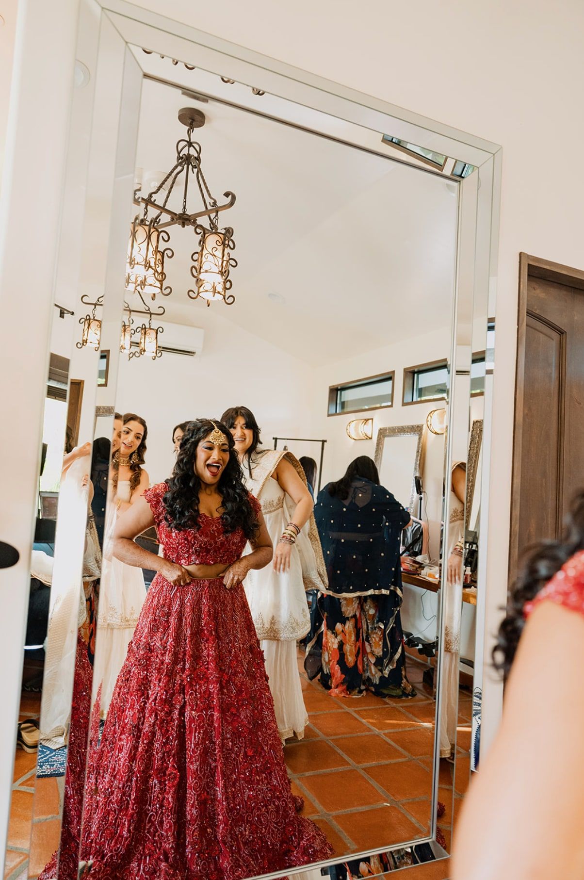 Bride in a red lehenga laughing at her reflection as her bridesmaids look on