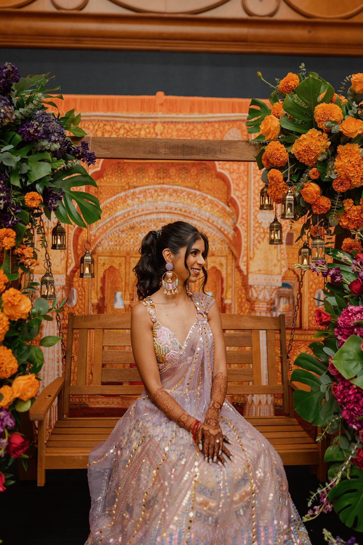 Bride in a blush lehenga smiling on a wooden swing framed by marigolds and hanging lanterns