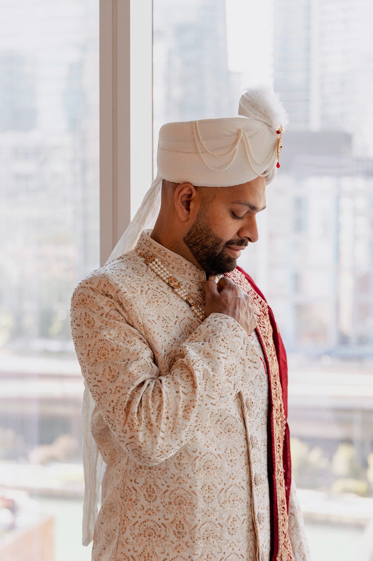Groom in an ivory sherwani and safa adjusting his collar by a tall window