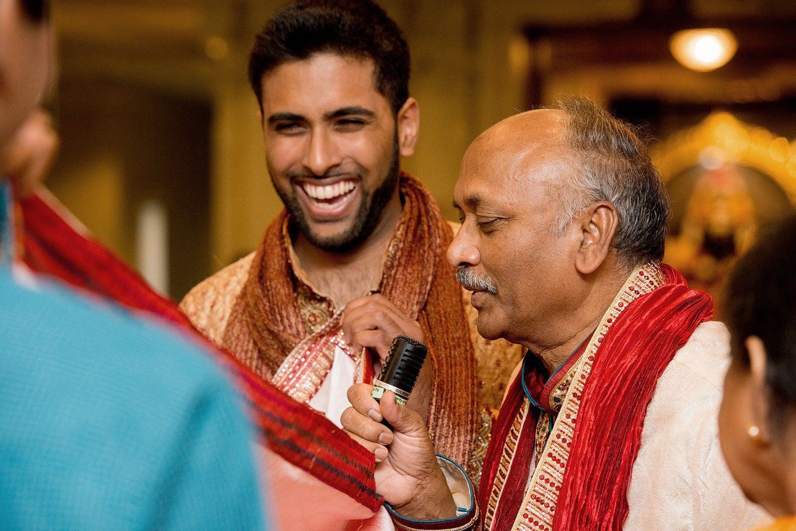 Groom laughing beside his father, who holds a microphone, in a red scarf