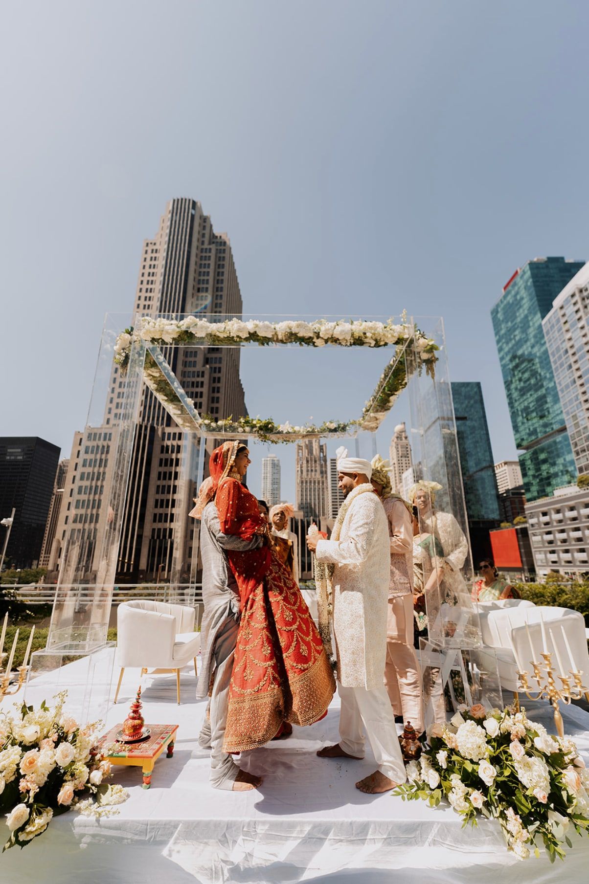 Couple facing each other under a clear mandap strung with white flowers, on a rooftop among skyscrapers