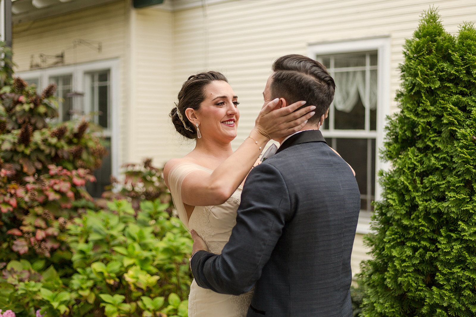A partner rests a hand on the other’s cheek, smiling, in front of a cream-colored house