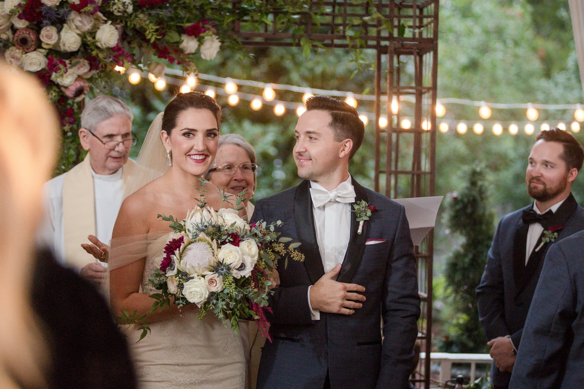 A couple smiles at the altar under string lights as the officiant speaks