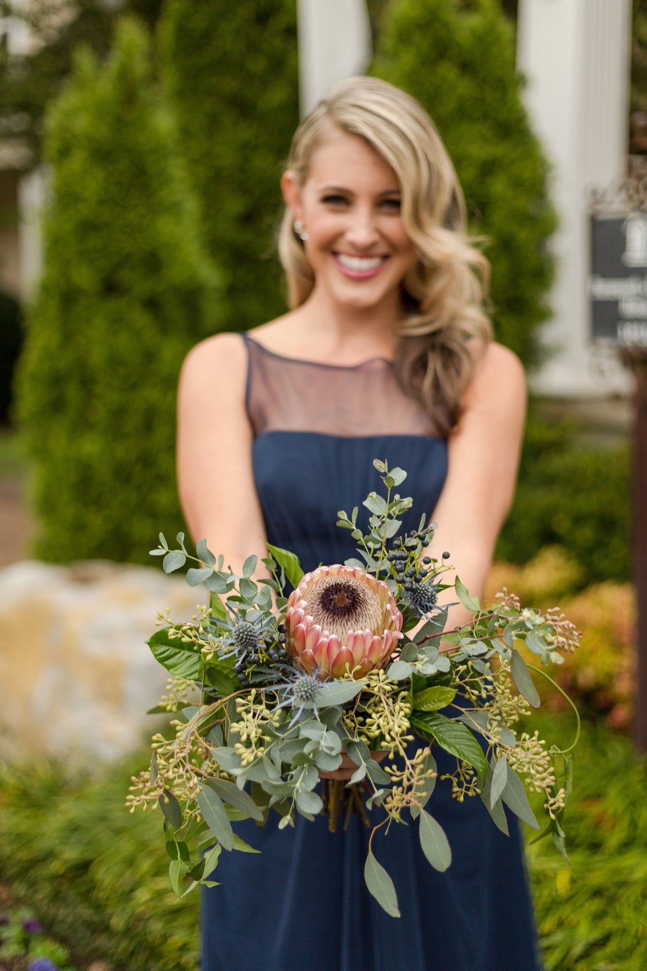 A bridesmaid in navy smiles, holding a bouquet with a pink protea