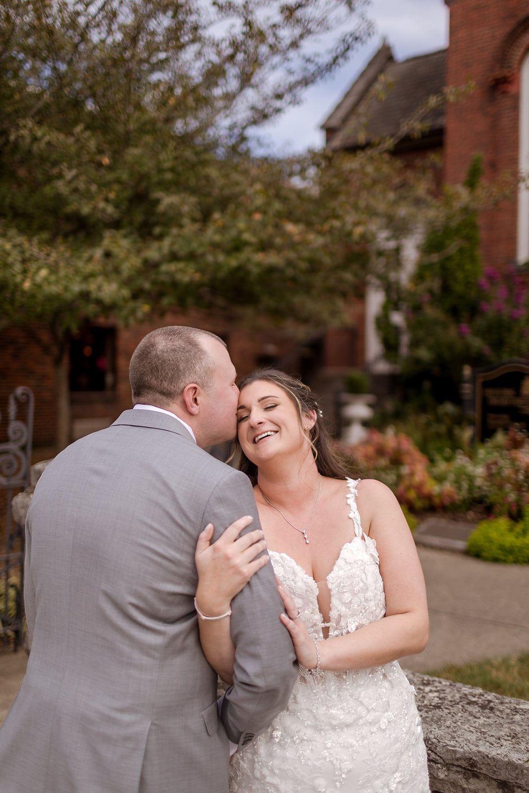 A couple shares a laughing embrace in a garden, red brick buildings behind them