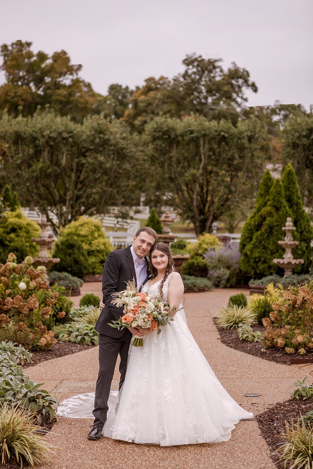 A couple stands on a garden path, a fountain and clipped hedges behind them