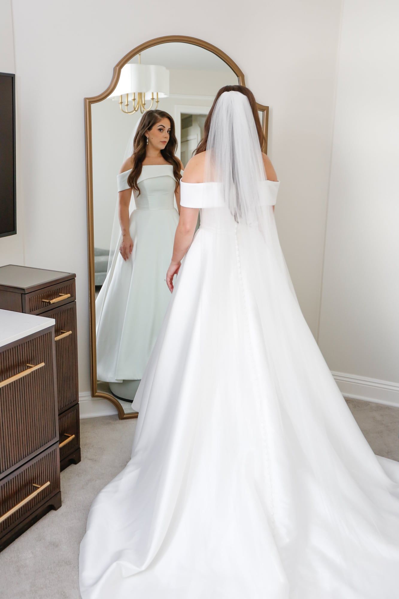 A long white gown seen from behind, facing a gold arched mirror while a bridesmaid watches