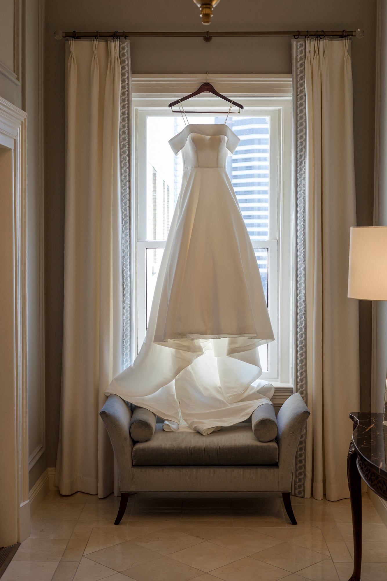 A wedding gown hangs in a sunlit window above a cushioned bench