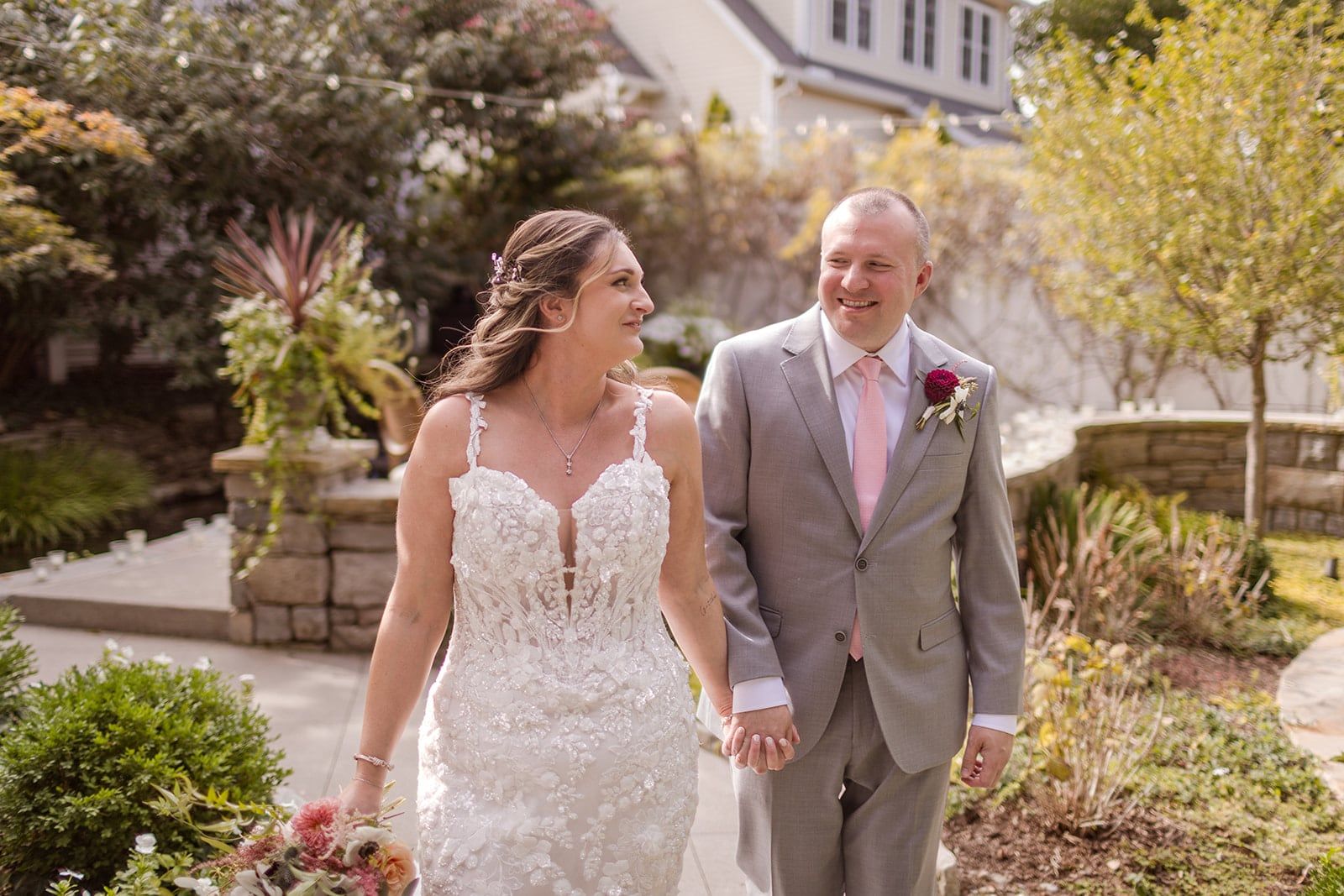 A couple walks hand in hand through a garden, smiling at each other