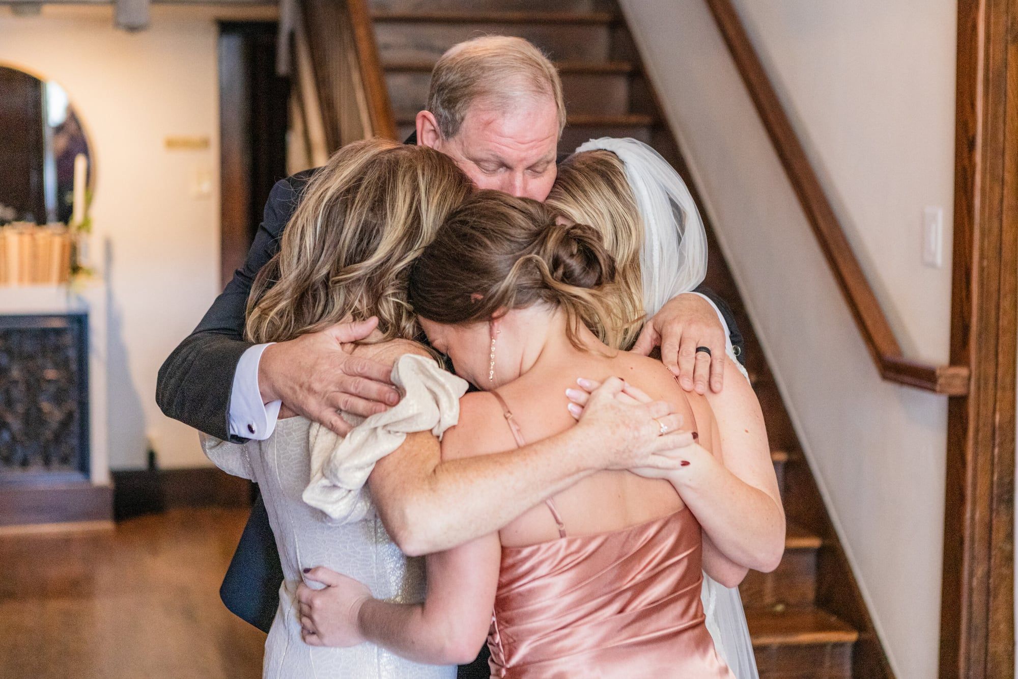 A father wraps his daughter, in her wedding dress, and a bridesmaid in pink in one big hug at the foot of the stairs
