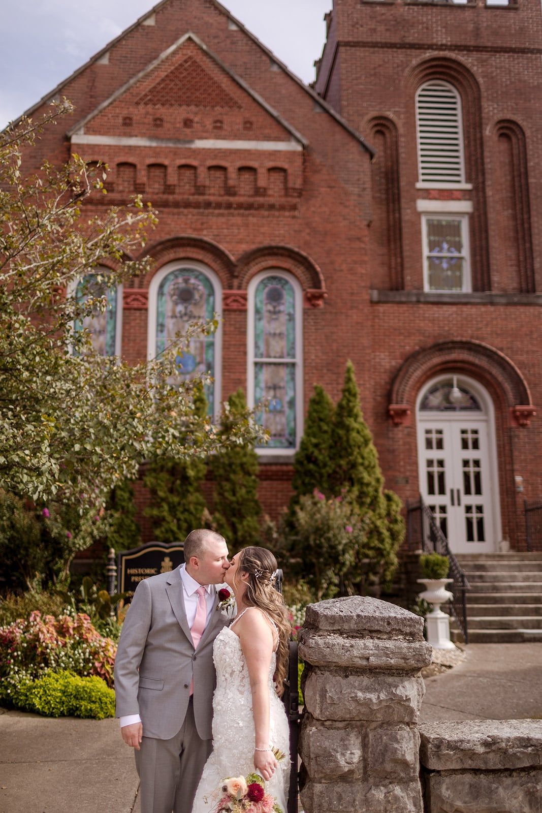A couple shares a kiss in front of a red brick church with tall arched windows