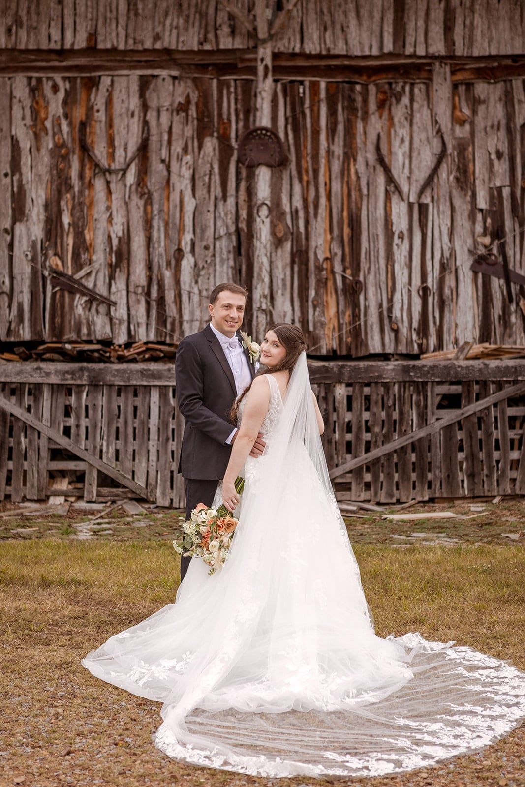 A couple in front of a weathered wooden barn, the long train of the dress spread across the grass