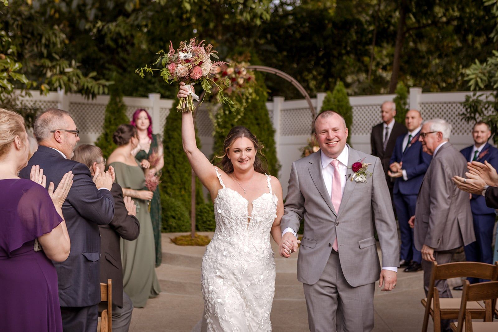 Newlyweds walk back down the aisle, one raising the bouquet high as guests applaud