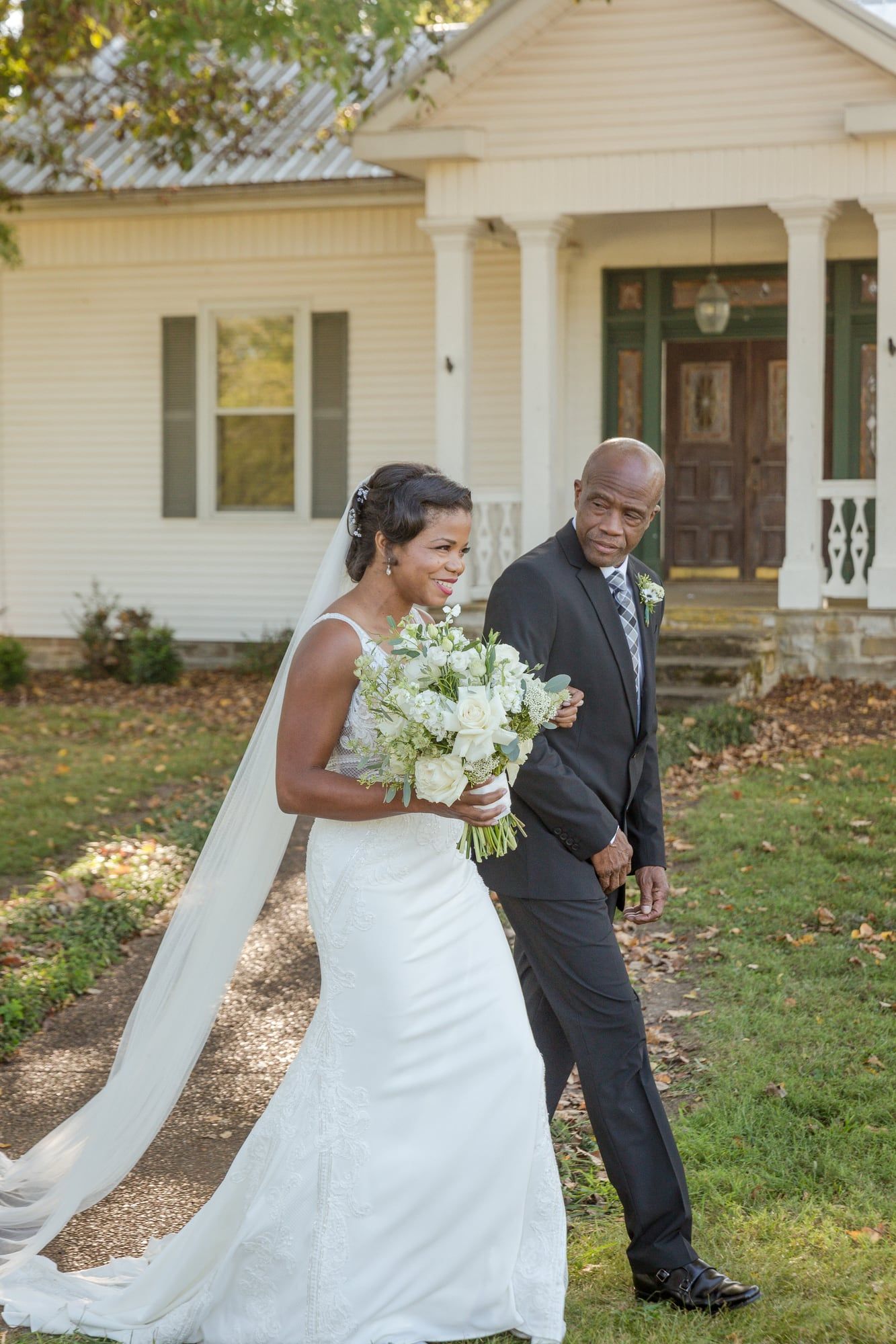 A bride in white walks across a lawn arm in arm with an older man in a dark suit, a white house behind them