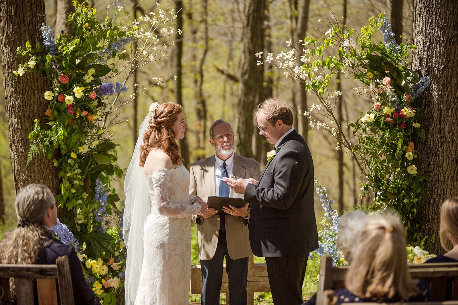 A couple faces each other at a woodland altar framed with flowers as the officiant reads