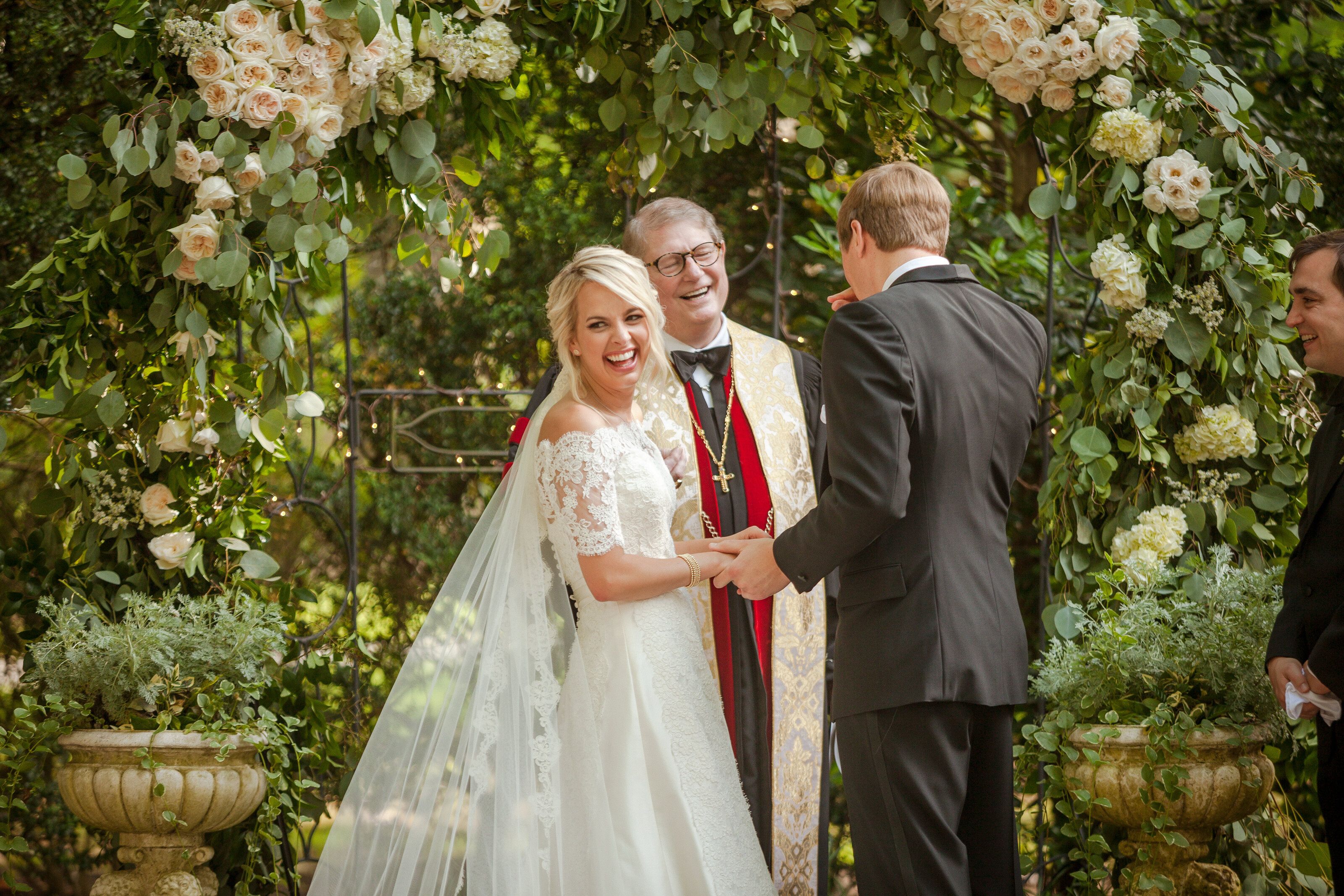 A couple holds hands and laughs through their vows under a floral arch while the officiant smiles