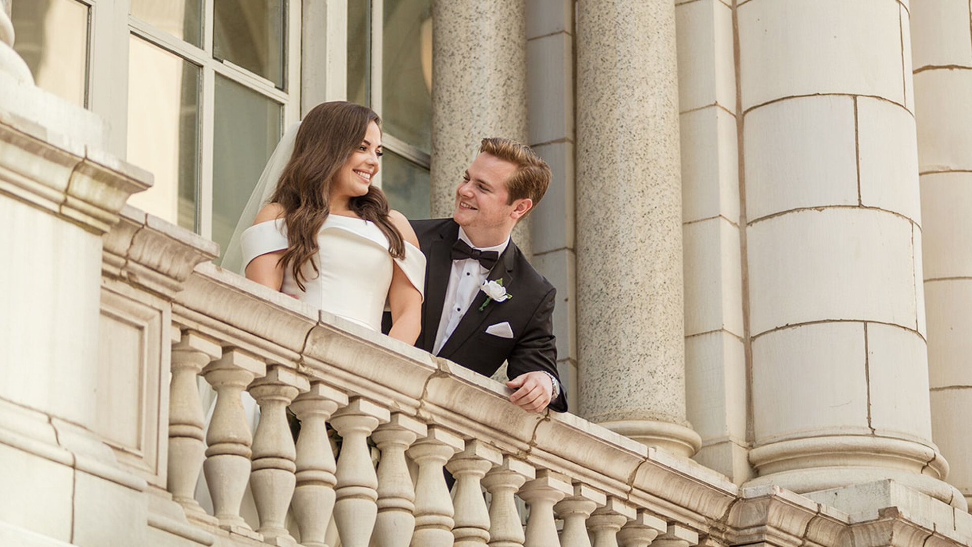 Shelby and Reid lean on a stone balustrade at The Hermitage Hotel, laughing together high above the street