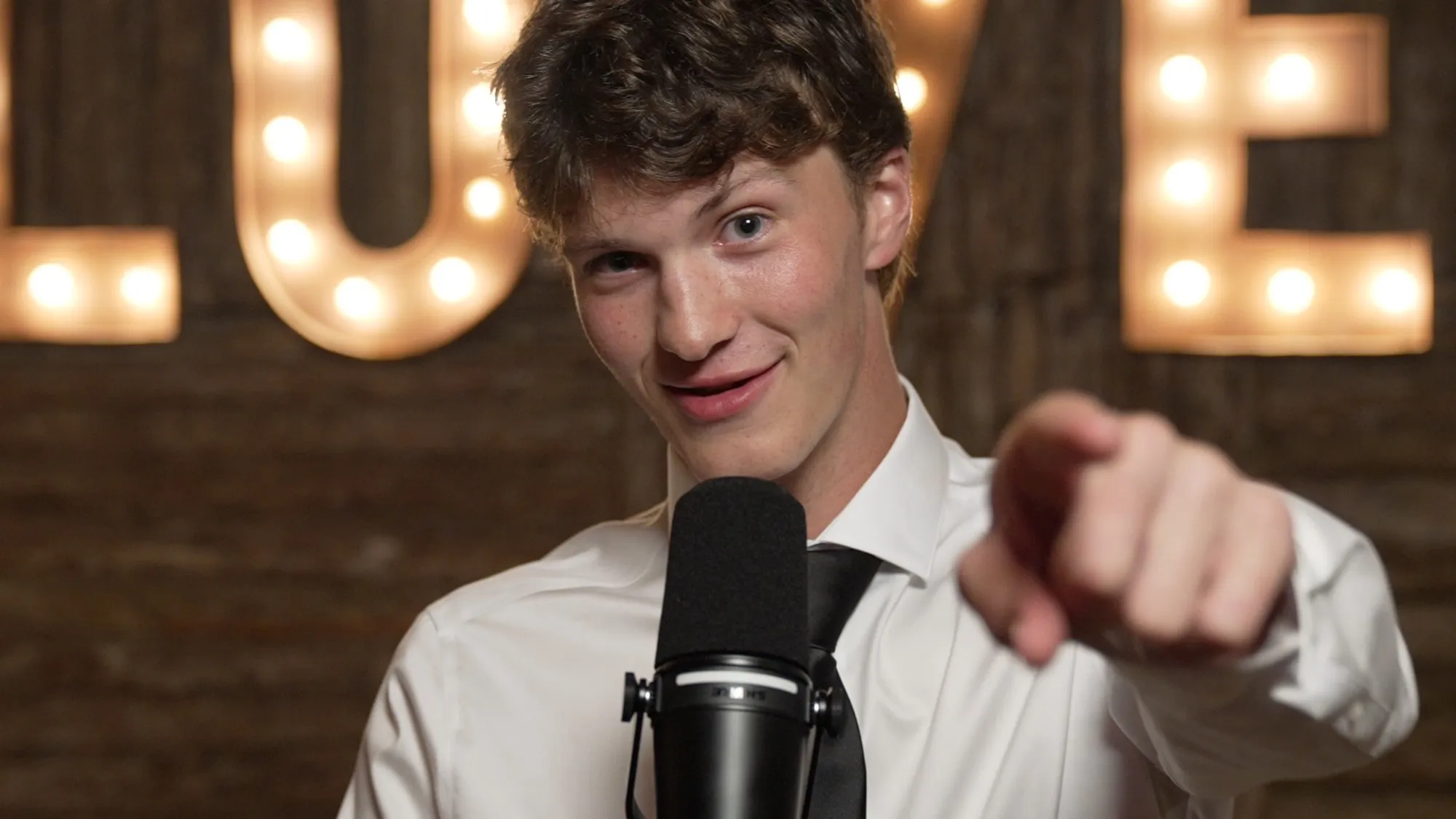 A wedding guest grins and points at the camera from the Unscripted booth mic, a lit LOVE marquee behind him