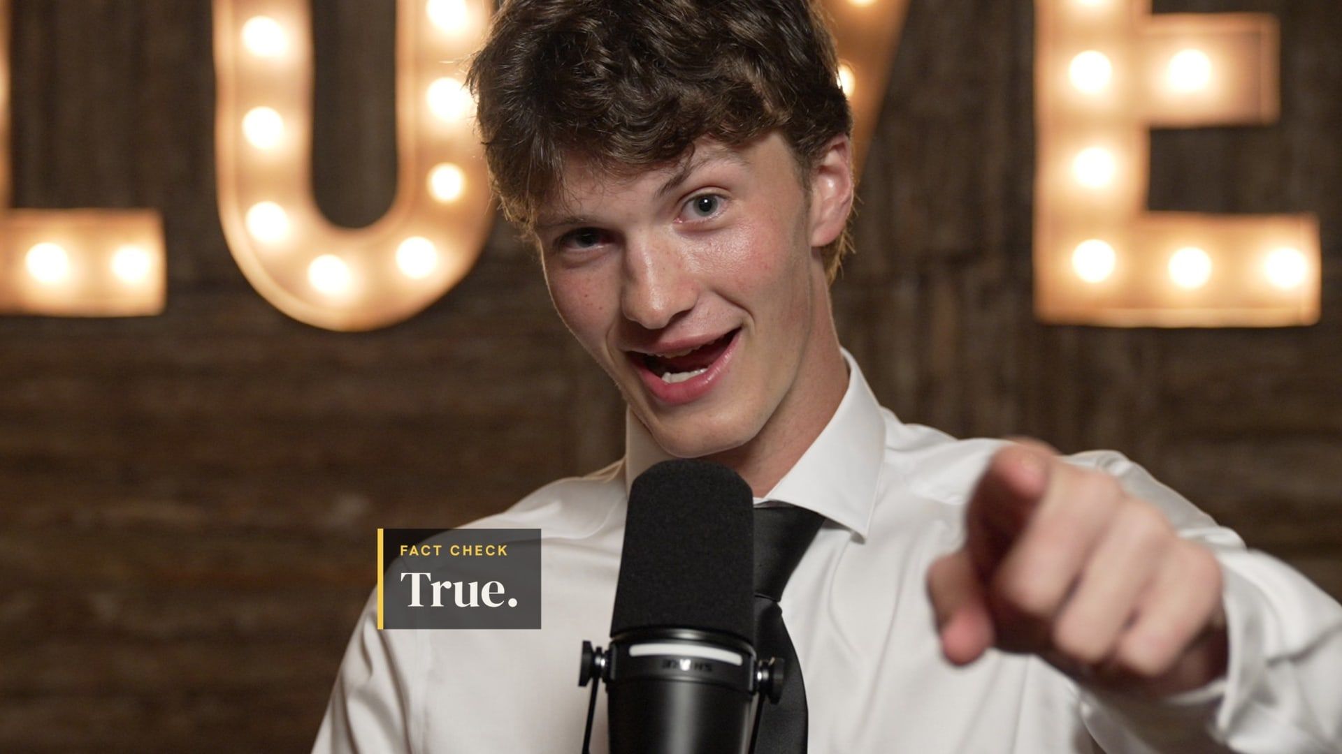 A guest grins and points at the camera from the Unscripted booth mic, a lit LOVE marquee sign behind, with an on-screen caption reading Fact check: True.