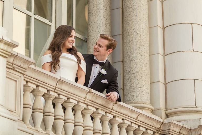 Shelby and Reid lean on a stone balustrade at The Hermitage Hotel, laughing together high above the street
