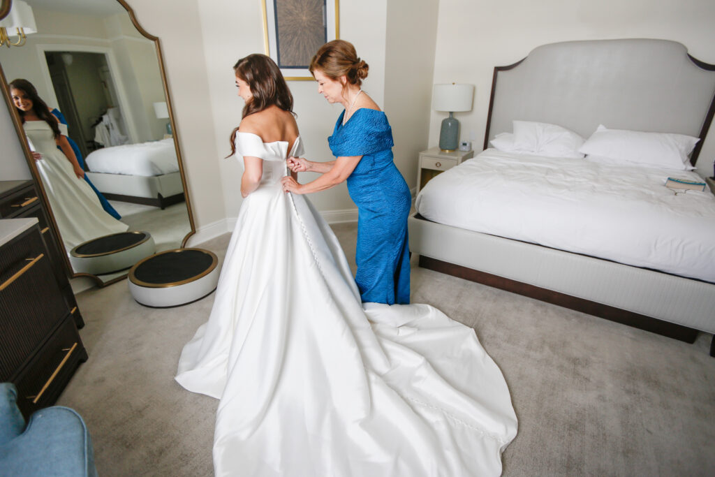 bride Shelby looks in a mirror as her mother zips up her wedding dress