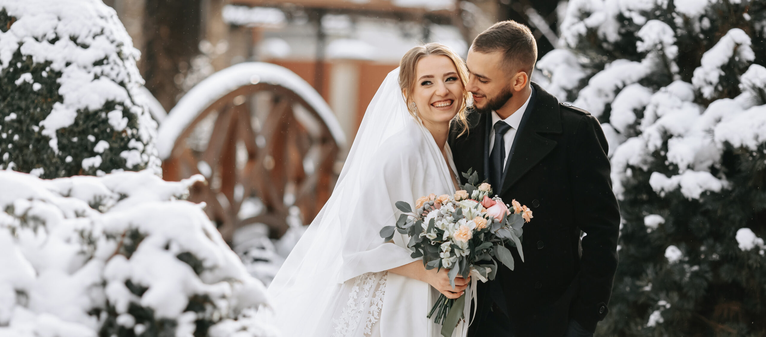 Beautiful and happy bride and groom are having fun among snowy trees. Bride and groom in the winter park. Bride with a bouquet of flowers in a wedding dress and poncho. Groom in a black coat.