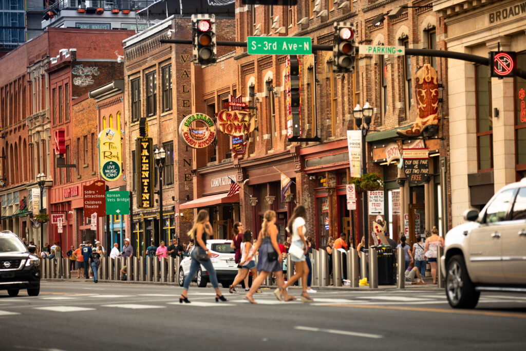 Bachelorette party group walking across Broadway in downtown Nashville during the day, with iconic neon signs and honky-tonk bars in the background.