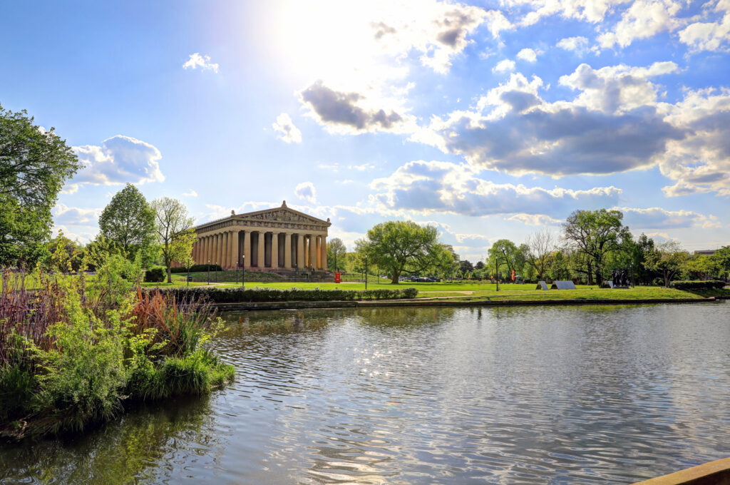 Sunlit view of The Parthenon in Centennial Park, Nashville, Tennessee, with a reflective pond and bright spring foliage — a grand and iconic location for romantic wedding portraits and elegant cinematic videography in the heart of the city.