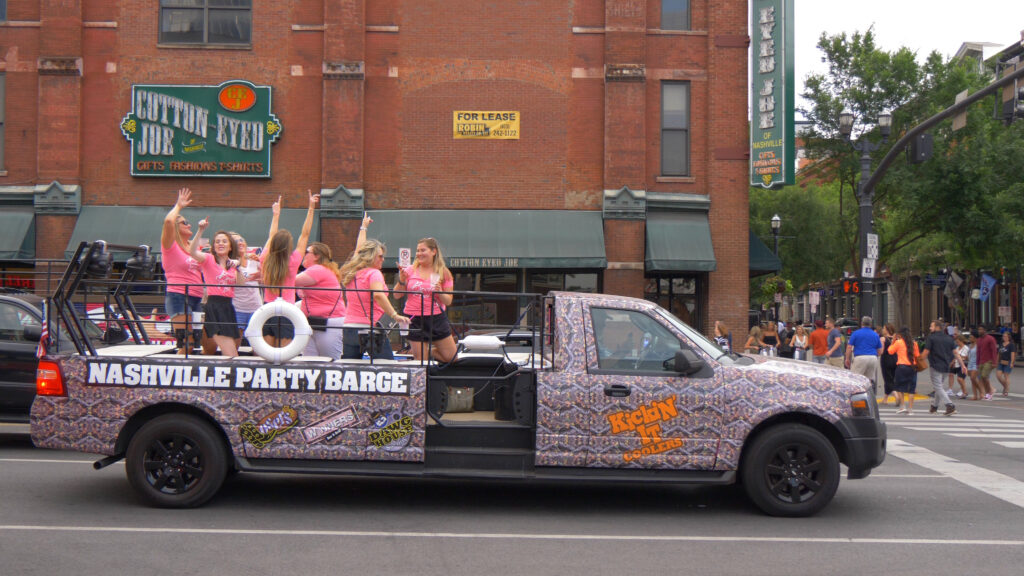 Group of women in matching pink shirts enjoying a bachelorette party on the Nashville Party Barge, cruising through downtown with Cotton Eyed Joe shop in the background. Perfect for fun-filled Nashville bachelorette weekends.
