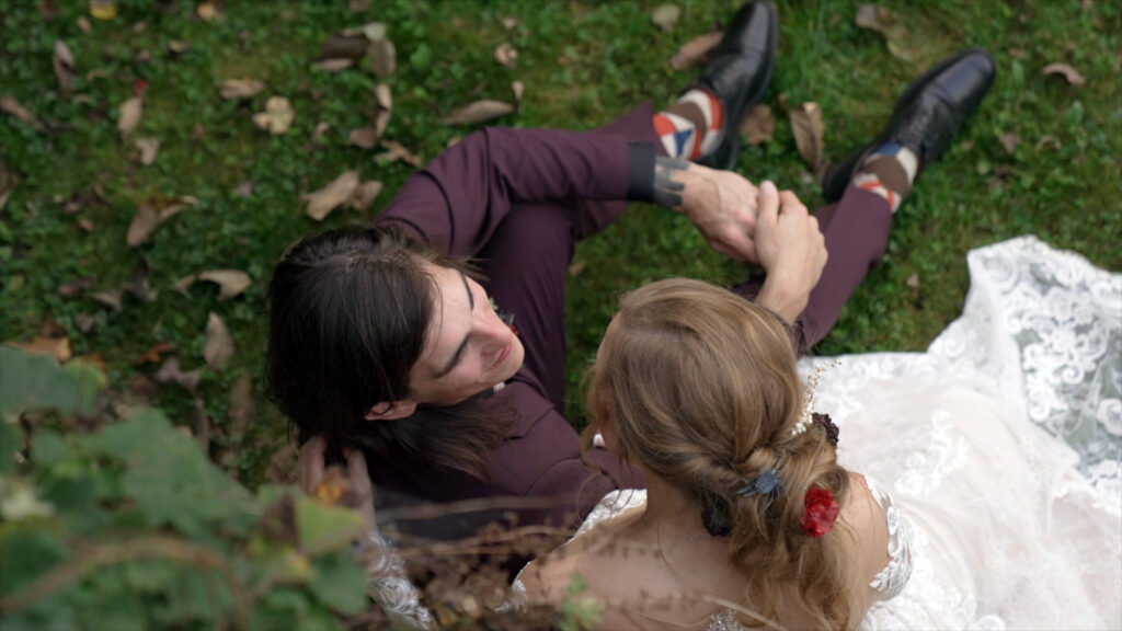 Bride and groom sitting together on the grass in an intimate moment at their Ruskin Cave wedding in Dickson, TN. Captured from above by With This Ring, Nashville wedding videographers, showcasing the bride’s lace gown and floral hairpiece during their fall celebration.