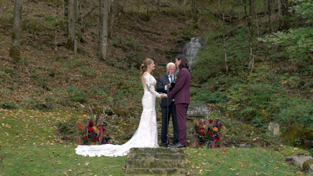 Bride and groom exchange vows in front of a cascading waterfall at Ruskin Cave in Dickson, TN, surrounded by vibrant fall florals. Filmed by With This Ring, Nashville wedding videographers specializing in cinematic outdoor weddings.